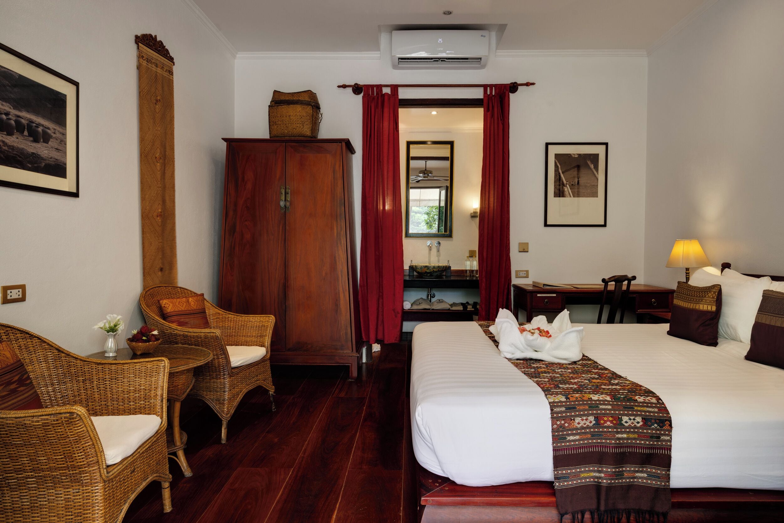 Une chambre d’hôtel comprenant un lit, deux fauteuils en osier, une armoire en bois, un bureau avec une lampe et un miroir.