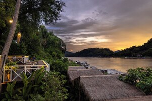 A riverside restaurant with outdoor seating overlooking a calm river, surrounded by lush greenery and a sunset.
