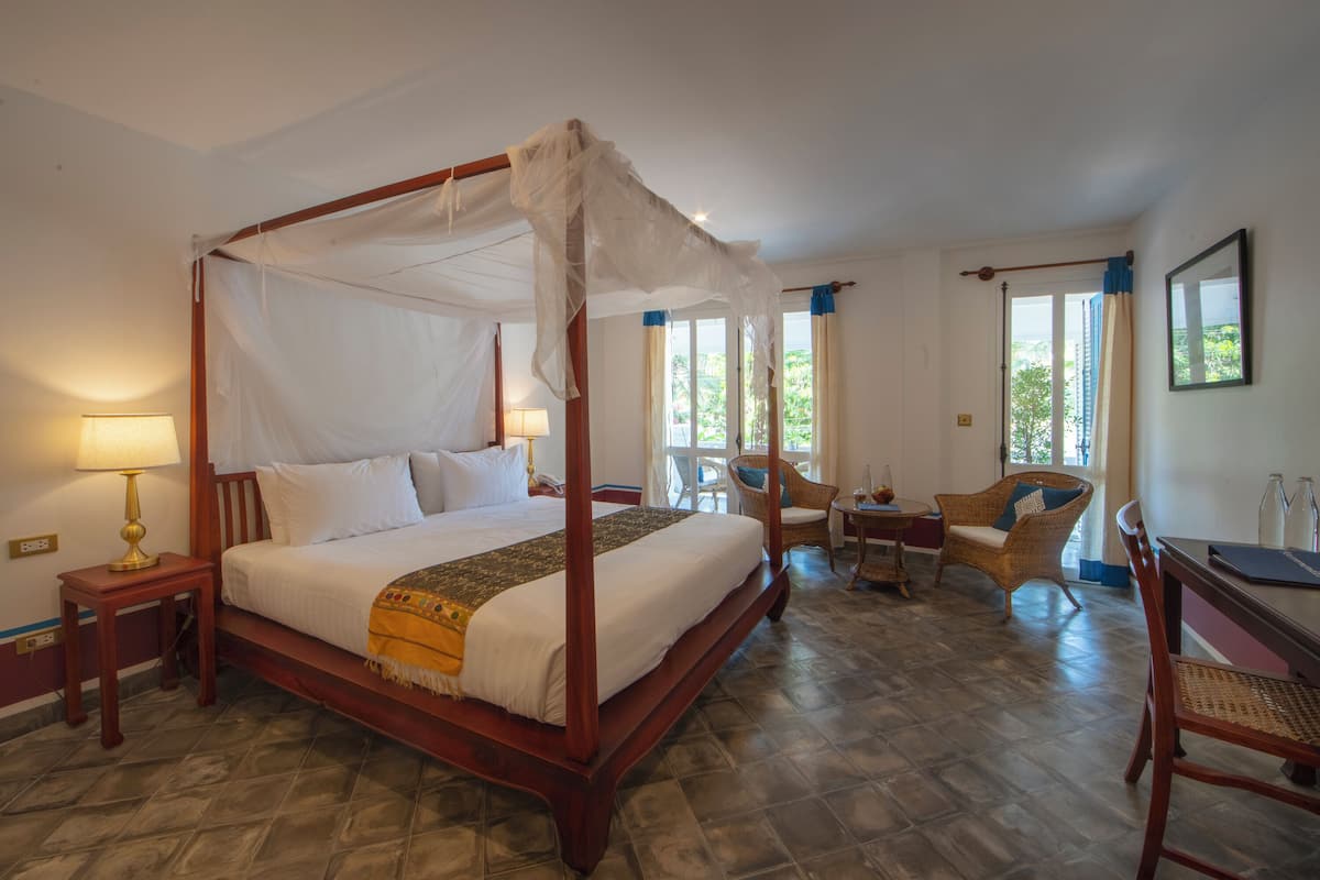 A four-poster bed with a canopy, a bedside table with a lamp, a wicker chair, and a desk with a chair.