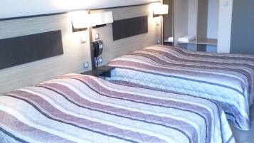 A hotel room with two beds, each with striped bedding, a bedside table with a lamp, and a wardrobe with hanging clothes.