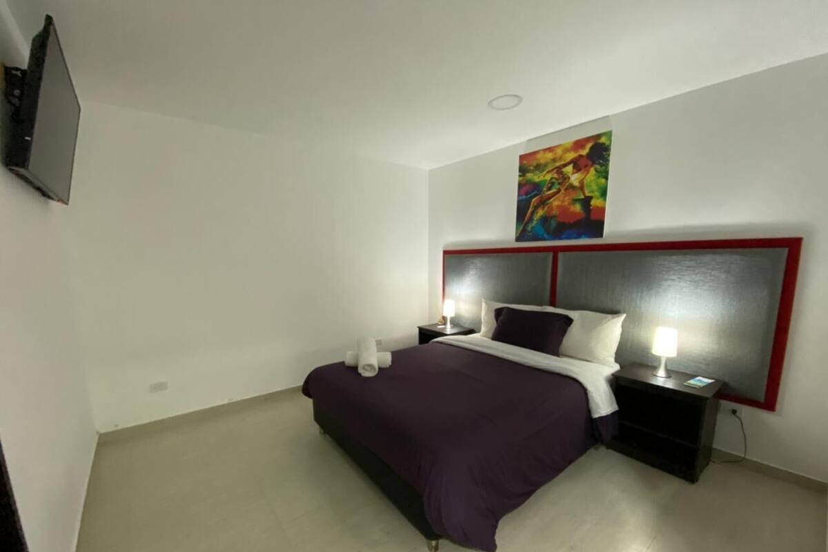 A bedroom with a bed, two bedside tables, a TV mounted on the wall, and a colorful painting above the bed.