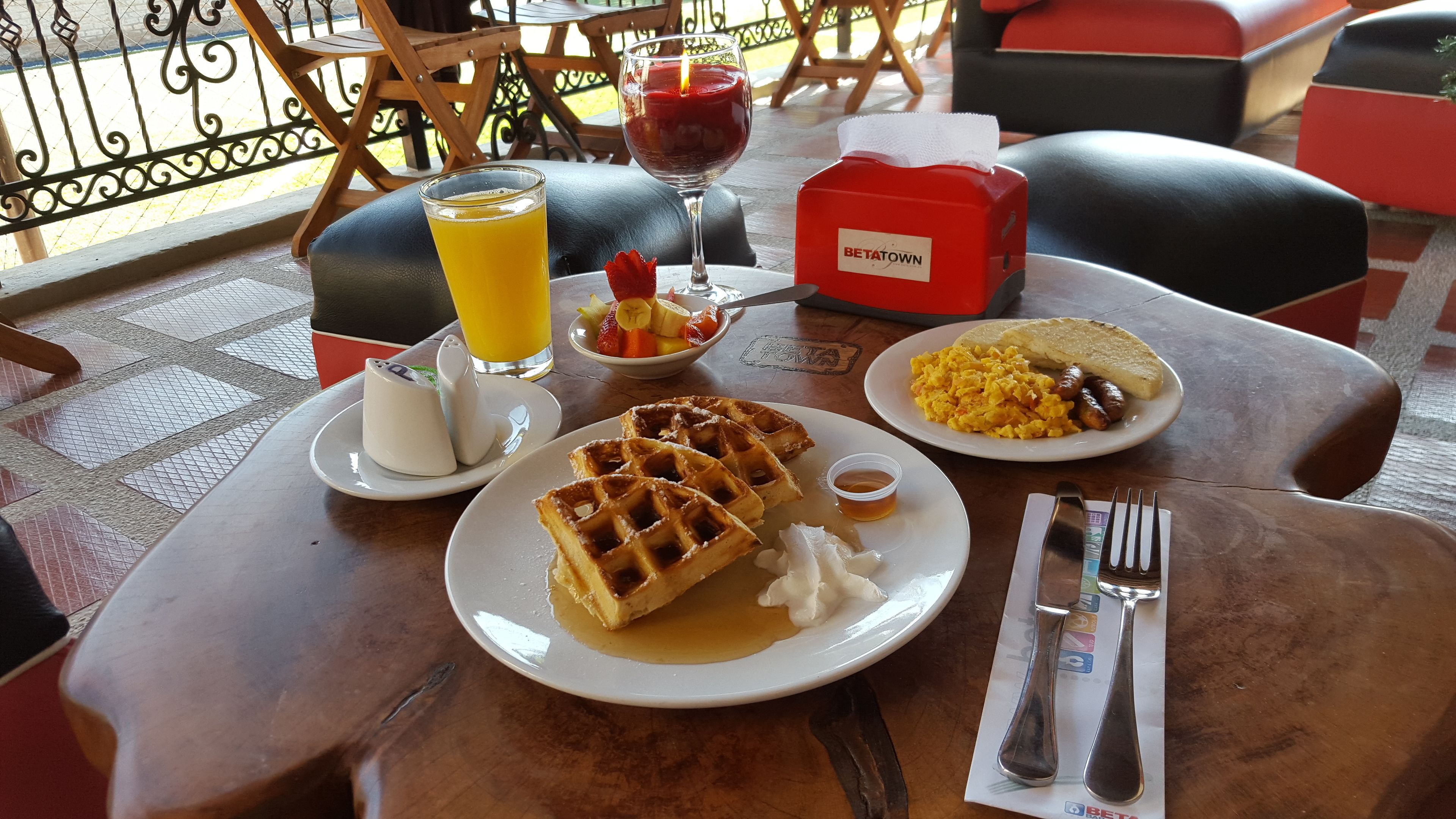 Desayuno con waffles, huevos revueltos, salchichas, tostadas, fruta y un vaso de jugo de naranja.