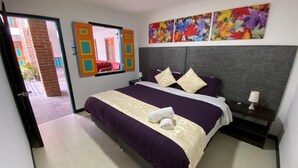 Habitación de hotel con una cama grande, una mesita de noche, un ventanal con vistas a un edificio de ladrillo y una puerta que da a un espacio exterior.
