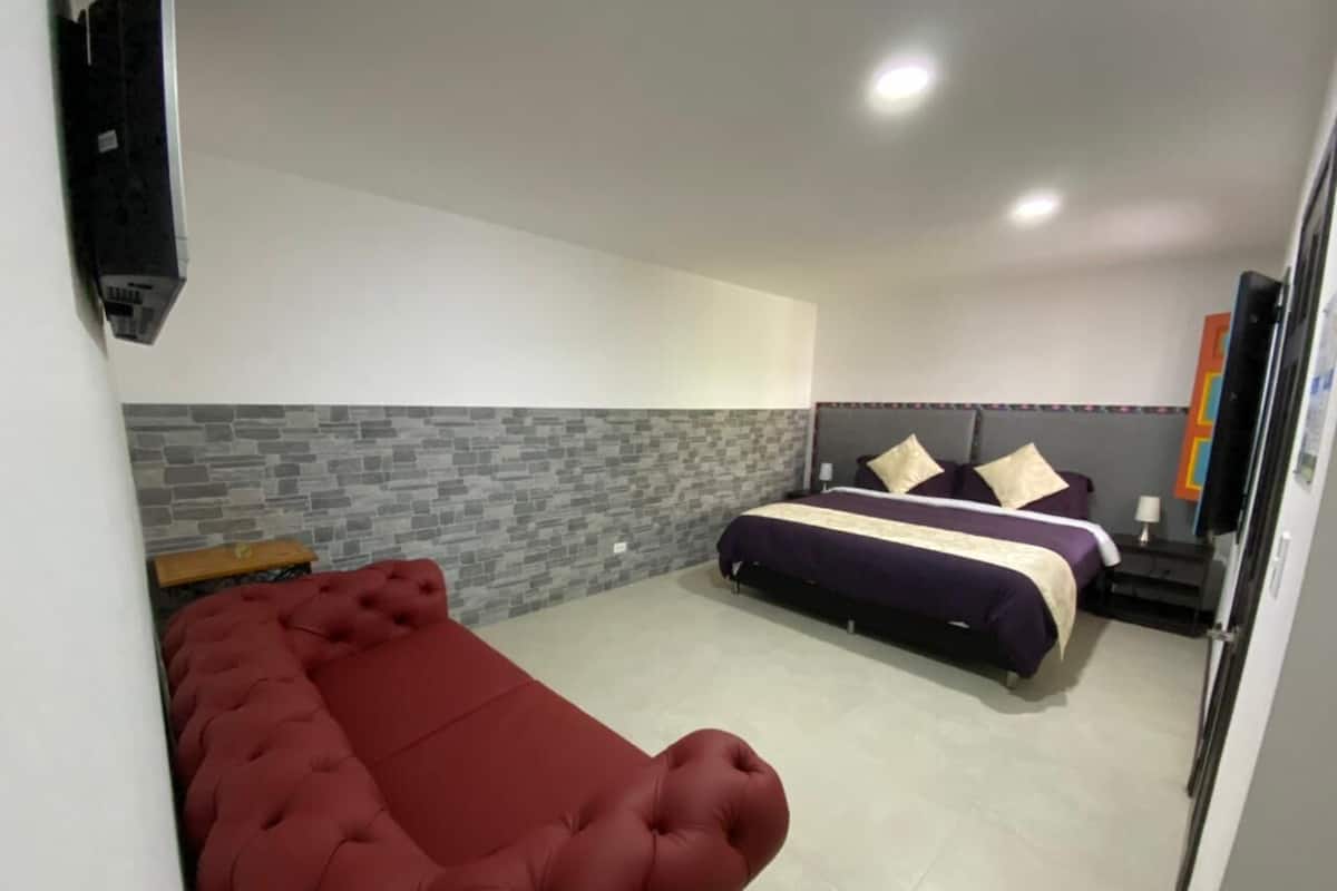A hotel room with a red sofa, a bed with a purple and white bedspread, a grey brick accent wall, and a white ceiling.