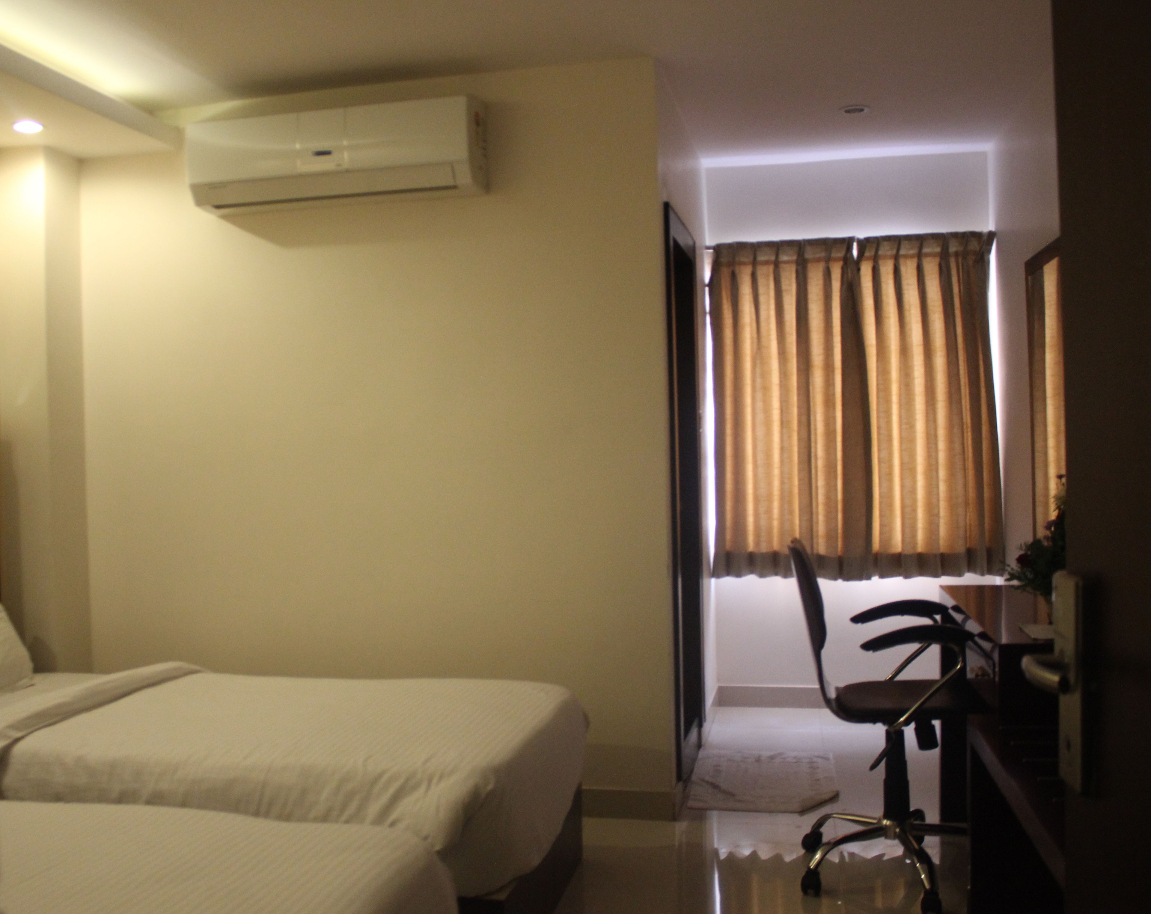 A hotel room with two beds, a desk with a chair, an air conditioning unit, and a window with curtains.