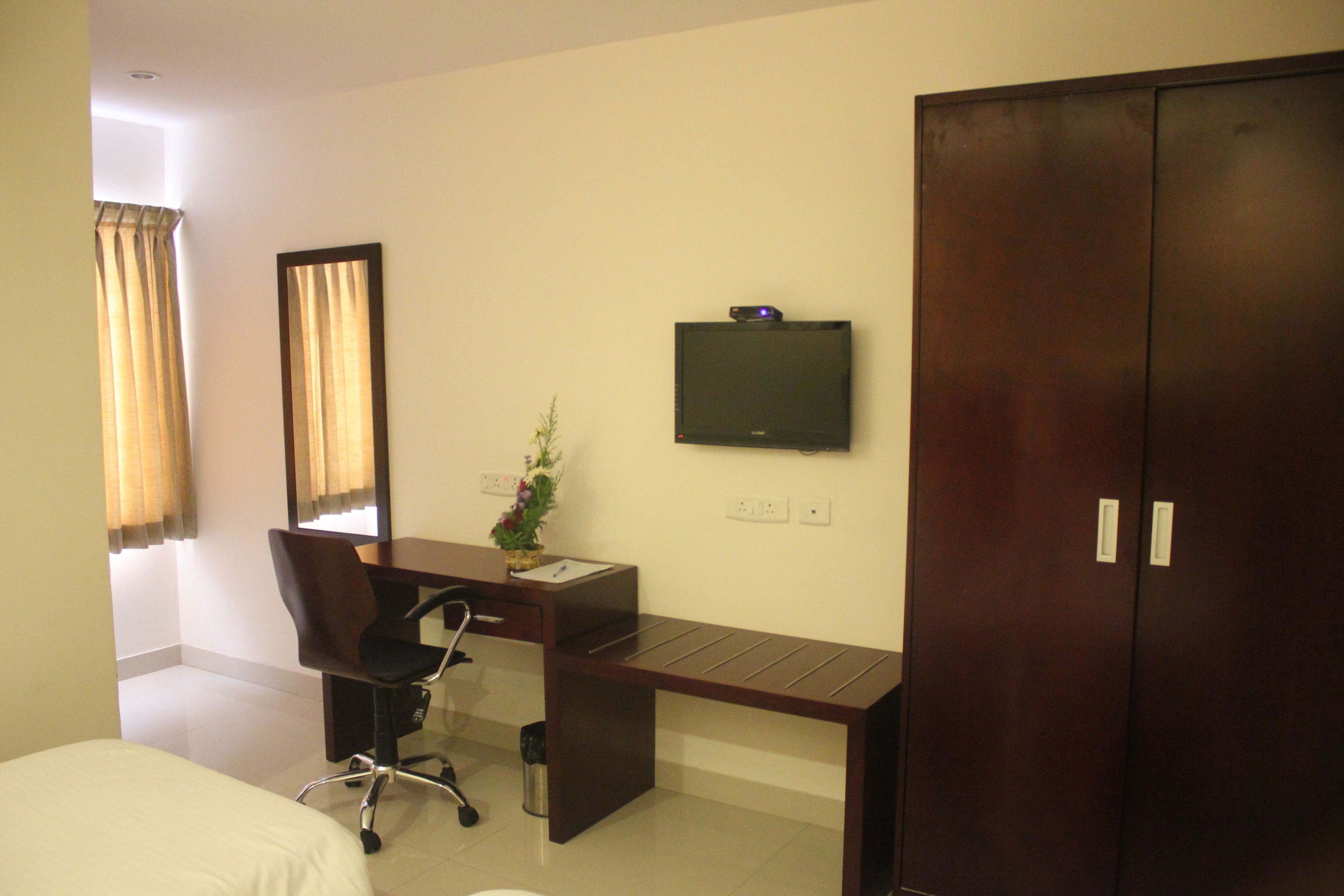 A hotel room with a bed, a desk with a chair, a television mounted on the wall, and a wardrobe.
