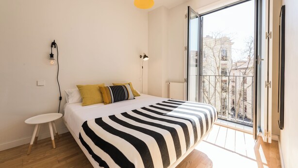 A bedroom with a large bed, a small bedside table, a wall-mounted lamp, and a sliding glass door to a balcony. - SmartRental Madrid Puerta del Sol (Madrid)