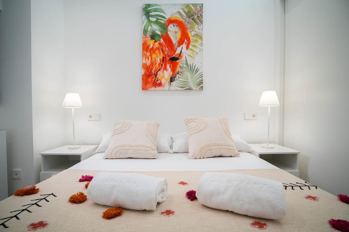 A hotel room with a large bed, two bedside tables with lamps, a tropical-themed painting on the wall, and towels on the bed.
