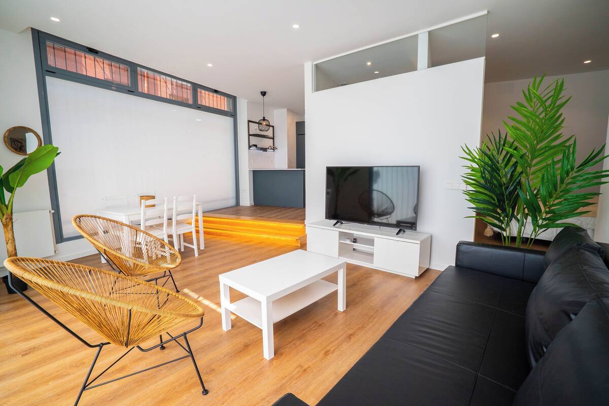 A modern living room with a sofa, white coffee table, wicker chairs, a TV, and a large plant.