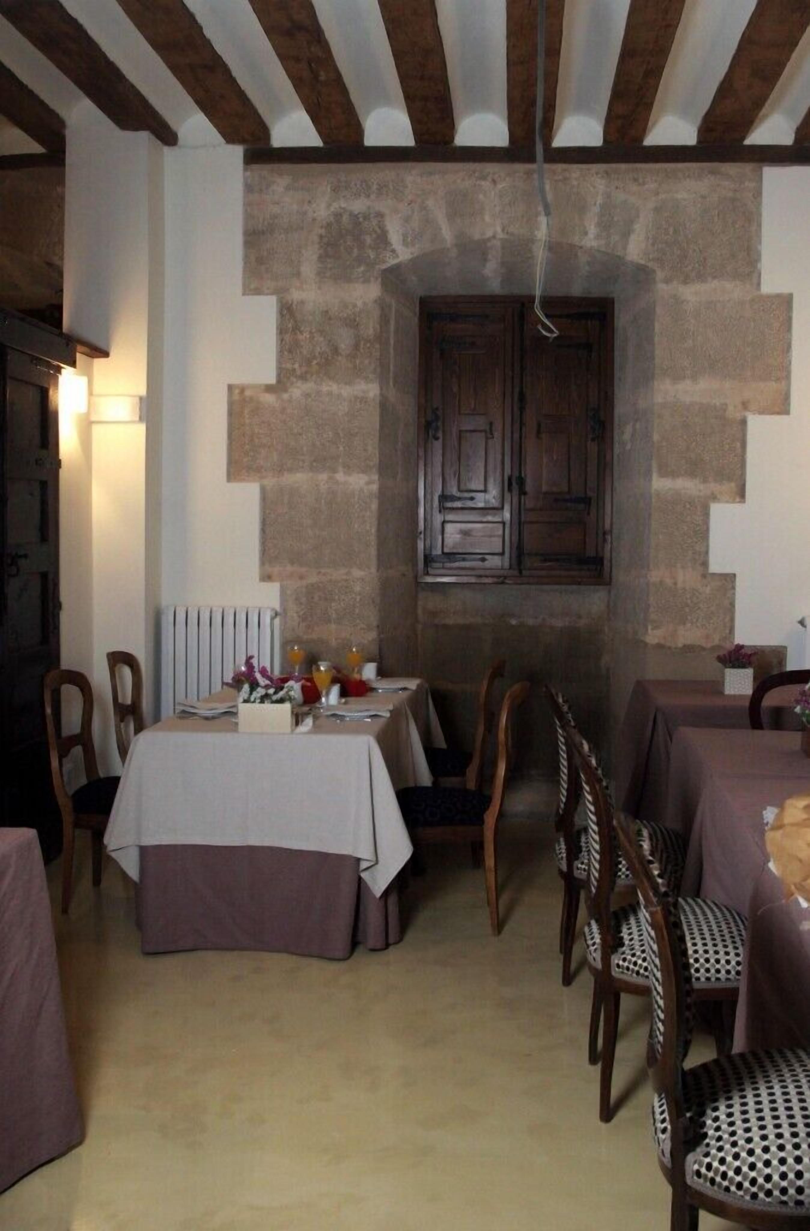 Uma área de jantar com parede de pedra, vigas de madeira no teto e uma porta de madeira rústica. Mesas montadas com toalhas brancas e cadeiras.