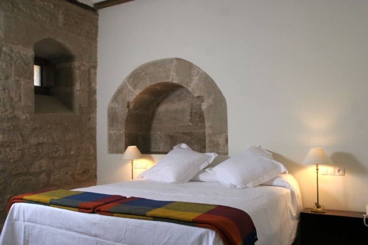 Ein Bett mit weißen Laken und einer bunten Decke, flankiert von zwei Nachttischlampen. Über dem Bett ist ein steinernes Rundbogenportal zu sehen.