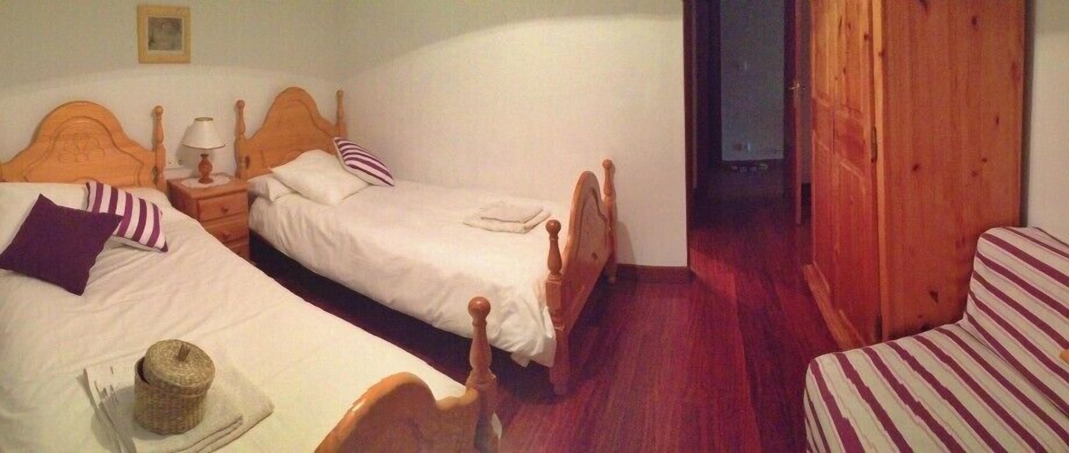 A room with two single beds, a wooden headboard, a nightstand with a lamp, a small table with a hat, and a striped chair.