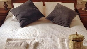 Ein ordentlich bezogenes Bett mit weißer Decke, zwei dunklen Kissen und einem kleinen gewebten Korb daneben.