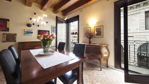 Un'area pranzo con un tavolo di legno, sedie e un vaso di fiori. Alle pareti ci sono dei dipinti e una finestra con vista su un edificio.