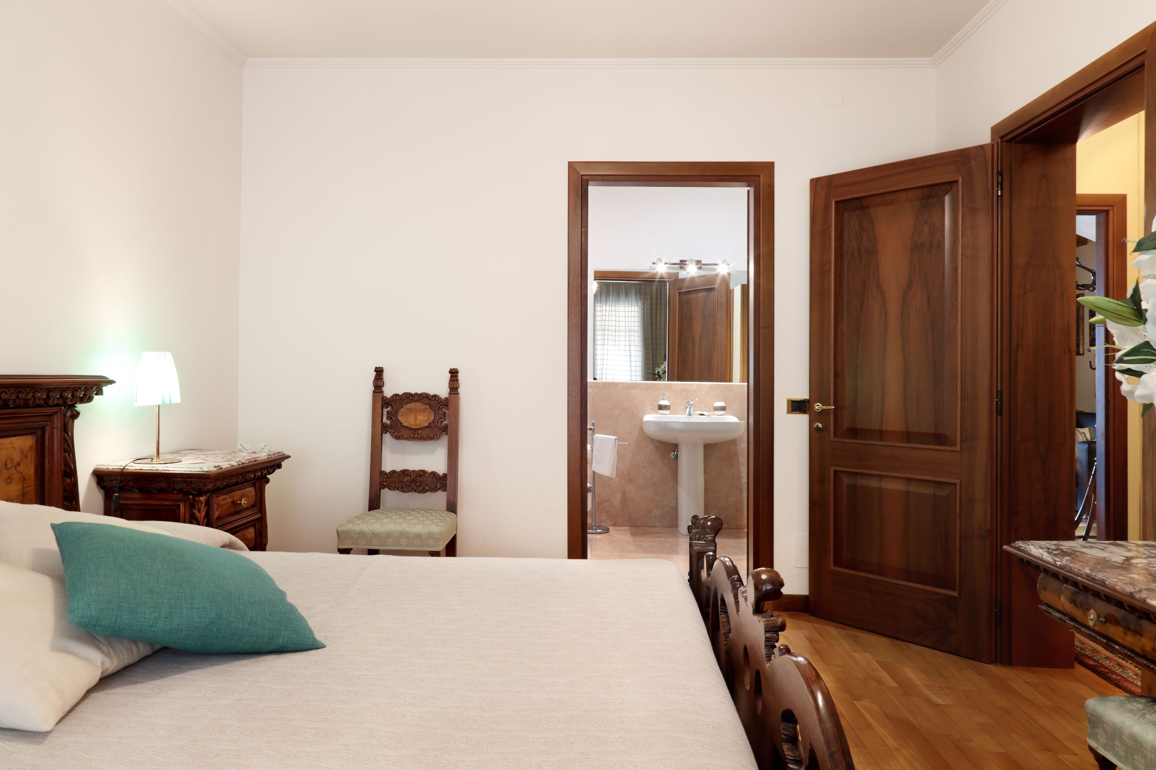 A hotel room with a bed, a wooden chair, a nightstand, a lamp, a wooden dresser, and a door leading to a bathroom.