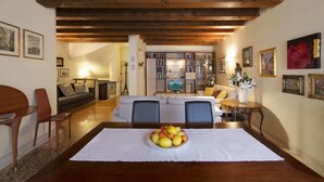 Un'area pranzo con tavolo in legno, sedie e una ciotola di frutta. Si vede anche un divano e una televisione sullo sfondo.