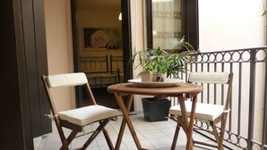 Un balcone con due sedie di legno, un tavolino rotondo, una pianta in vaso e vista sull'interno attraverso una porta aperta.
