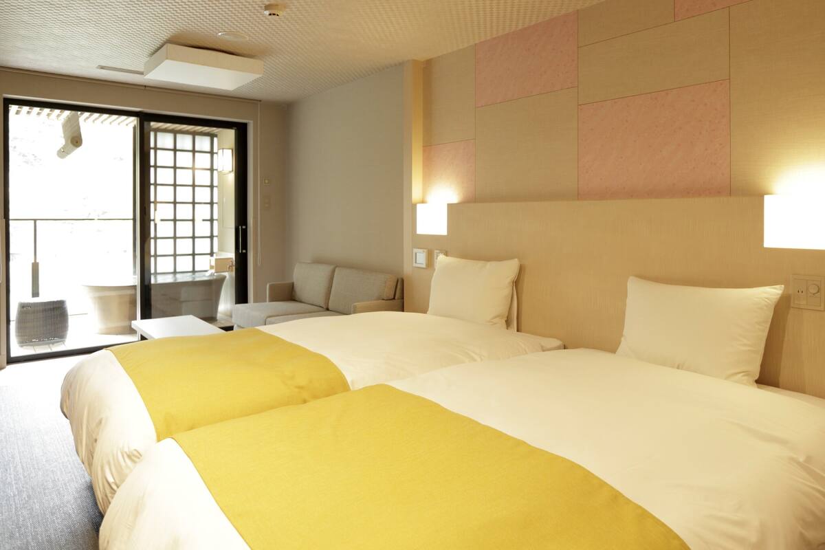 A hotel room with two beds, a sofa, a small table, and a view through a window.