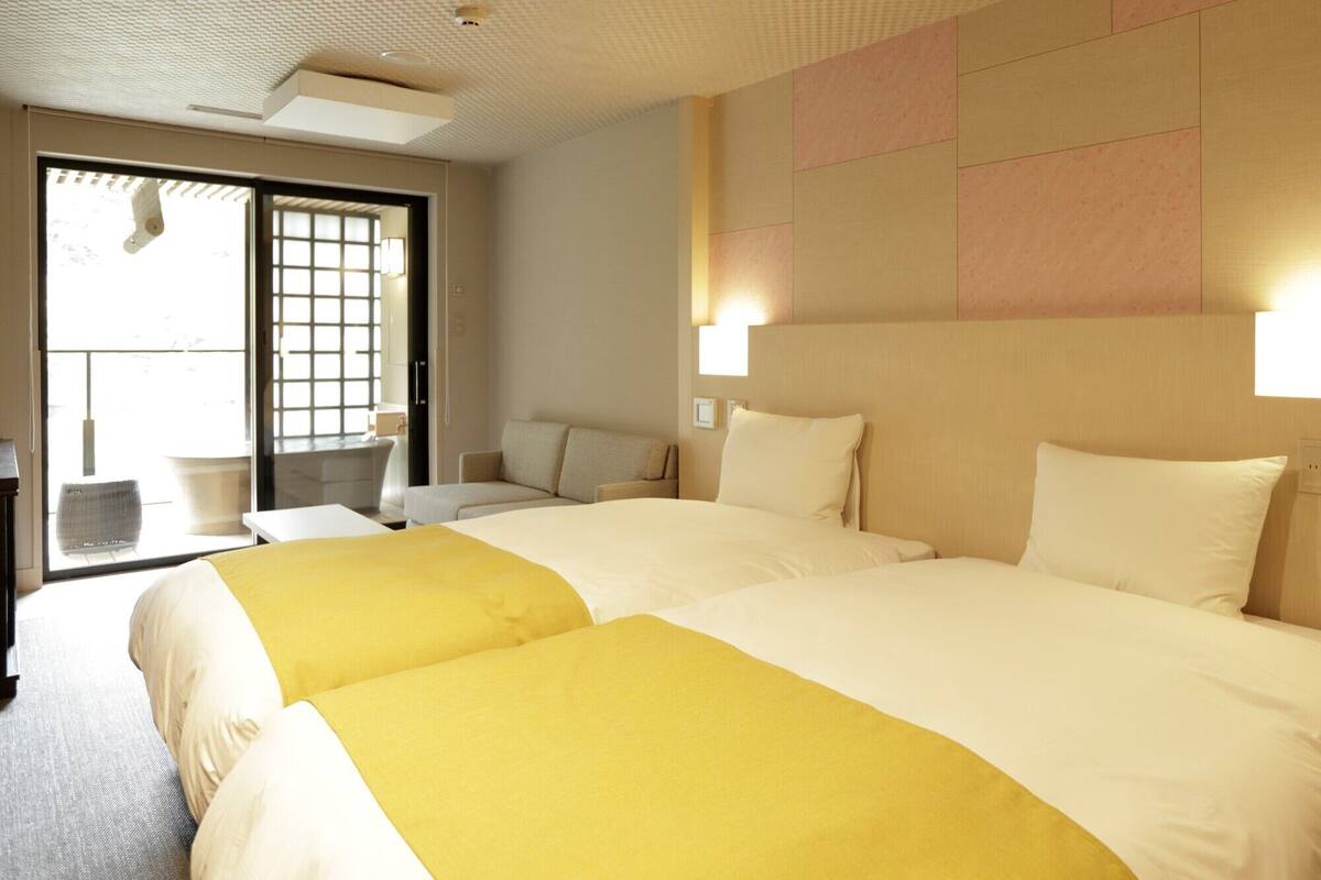 A hotel room with two beds, a sofa, a small table, and a view of the outside through a sliding door.