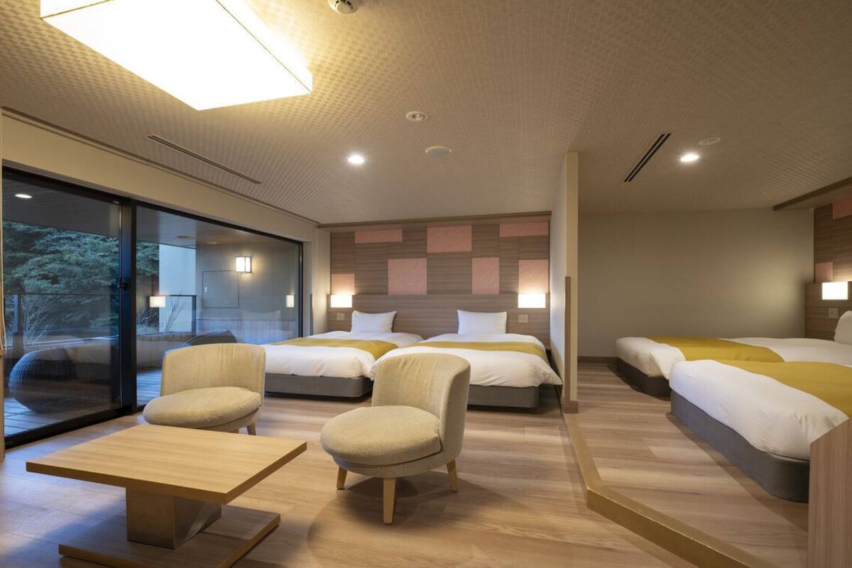 A modern hotel room with a large sliding glass door, two beds with yellow bedding, a small sofa, and a wooden coffee table.