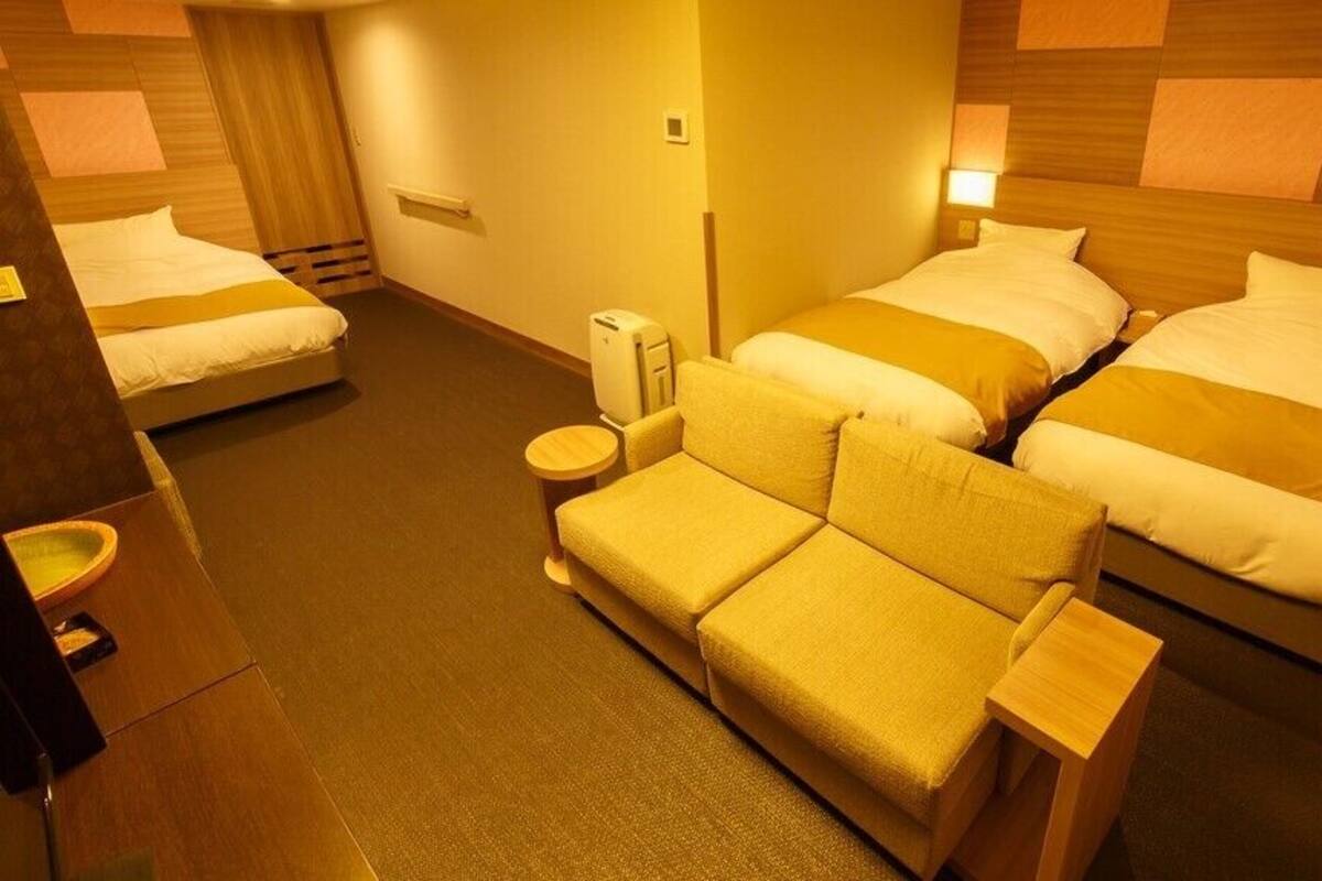 A hotel room with two beds, a sofa, a bedside table, a lamp, and a wall-mounted heater.