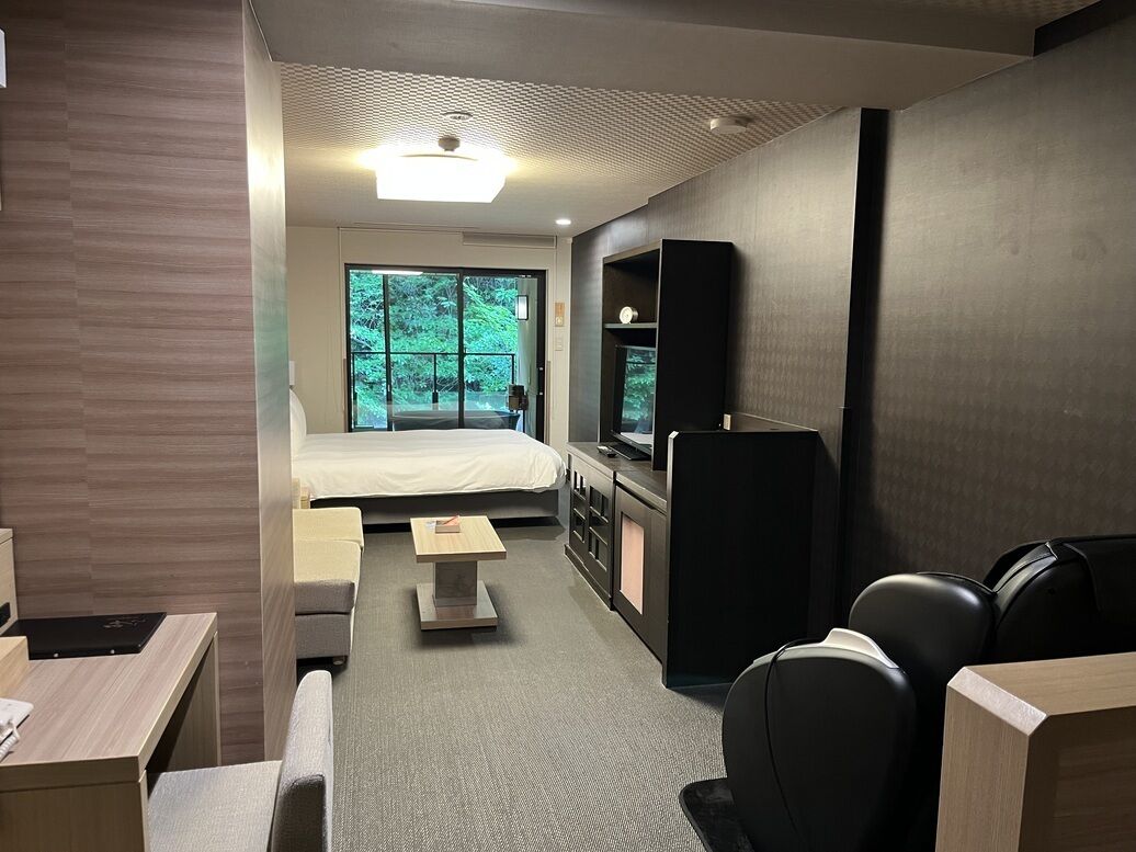 Ein modernes Hotelzimmer mit Bett, Schreibtisch, Sessel, Fernseher und Blick auf Grünfläche.
