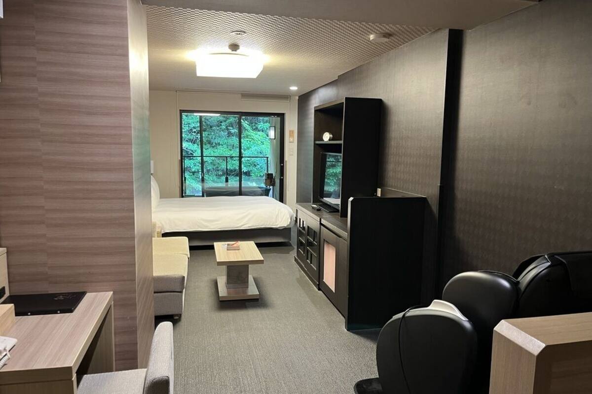 A modern hotel room with a bed, a desk, a chair, a TV, and a view of greenery outside.