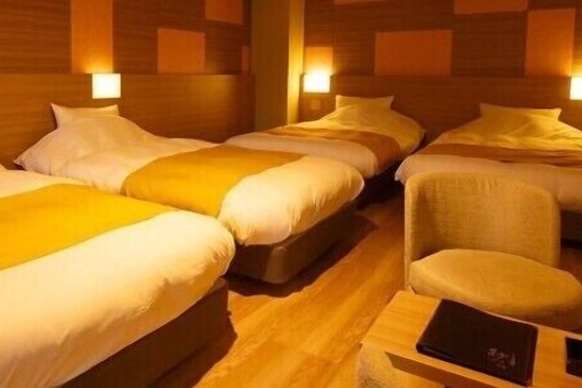 A hotel room with three beds, a wooden floor, and wall-mounted lamps.