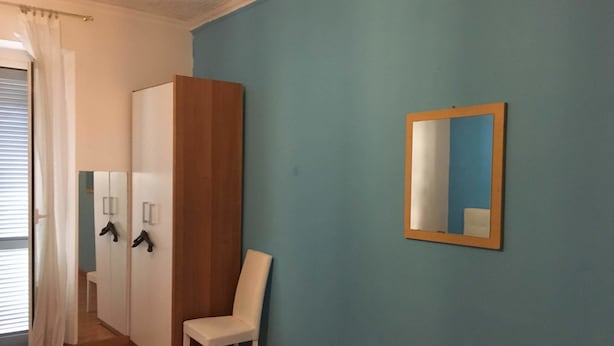 A room with a wooden wardrobe, a white chair, a mirror on the wall, and a window with white curtains. - Appartamenti Soperga (Milan)