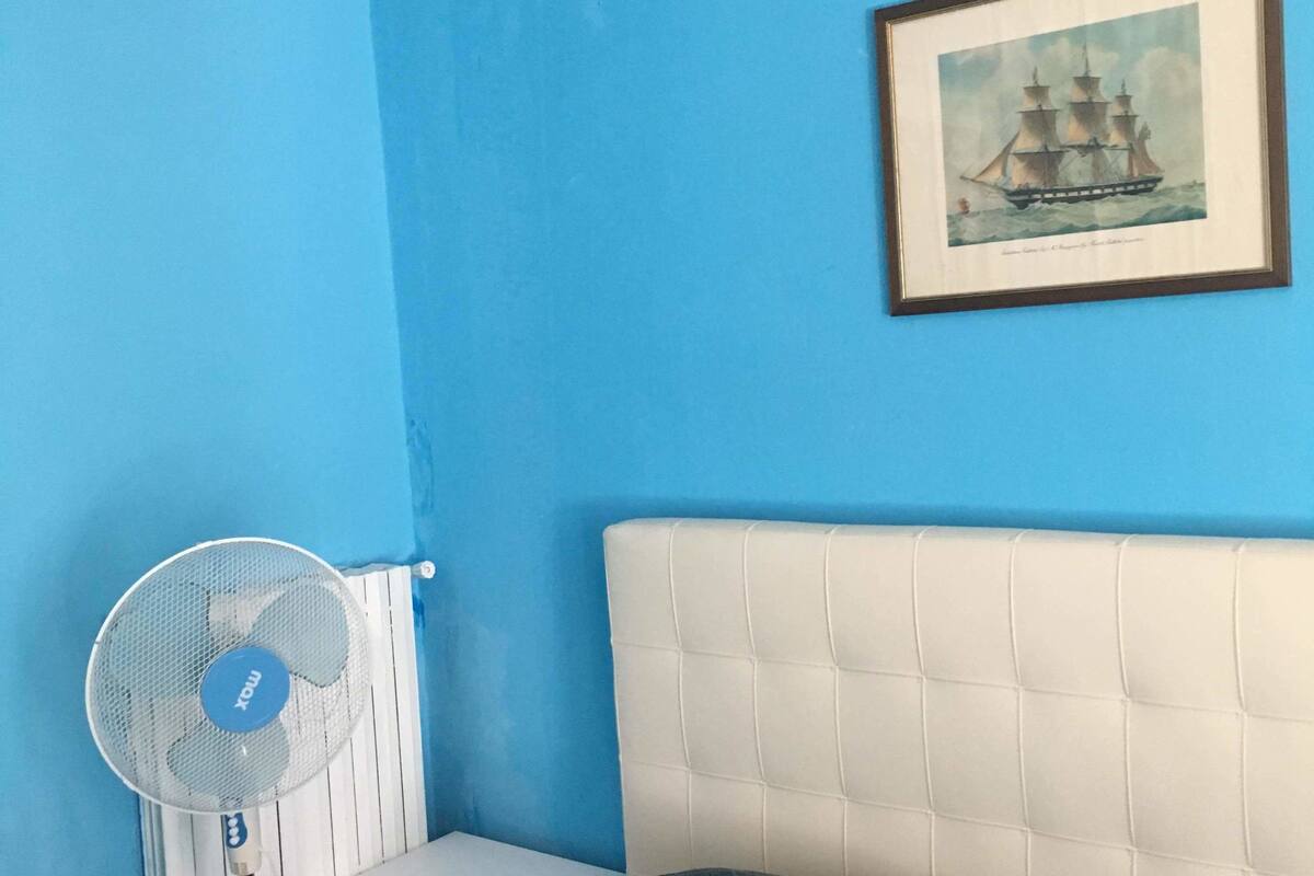 Una habitación con pared azul, un cuadro enmarcado de un barco, cabecera blanca y un ventilador de pie.