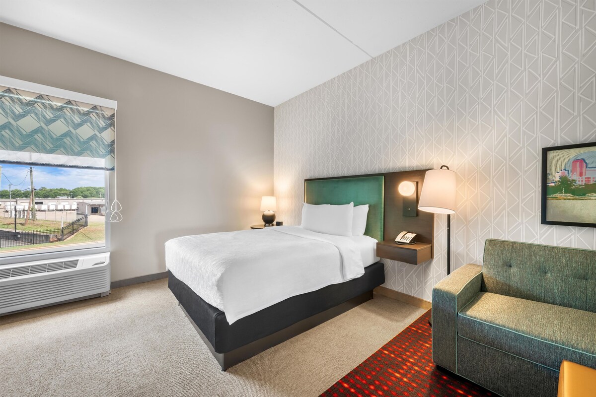 A hotel room with a bed, a nightstand, a lamp, a sofa, a window with a view, and a patterned wall.