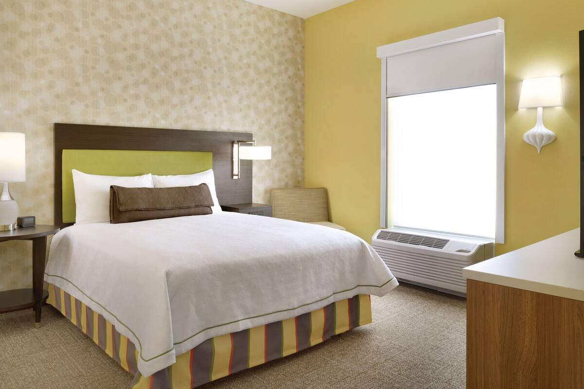 Une chambre d’hôtel comprenant un lit, une table de chevet, une lampe, une fenêtre avec des stores et un climatiseur fixé au mur.