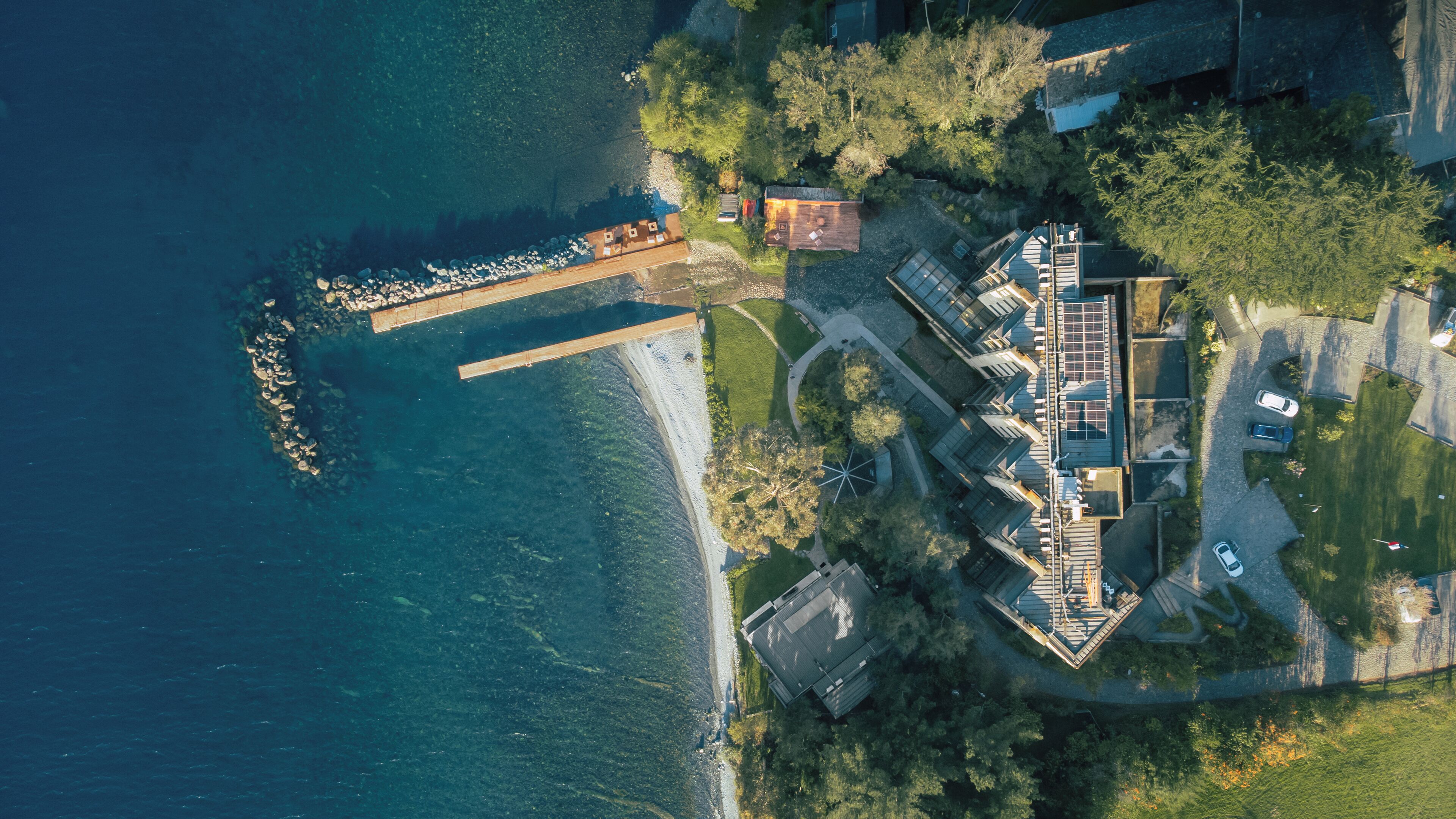 An aerial view of a coastal property with a dock, a large house with a sloped roof, and surrounding greenery.