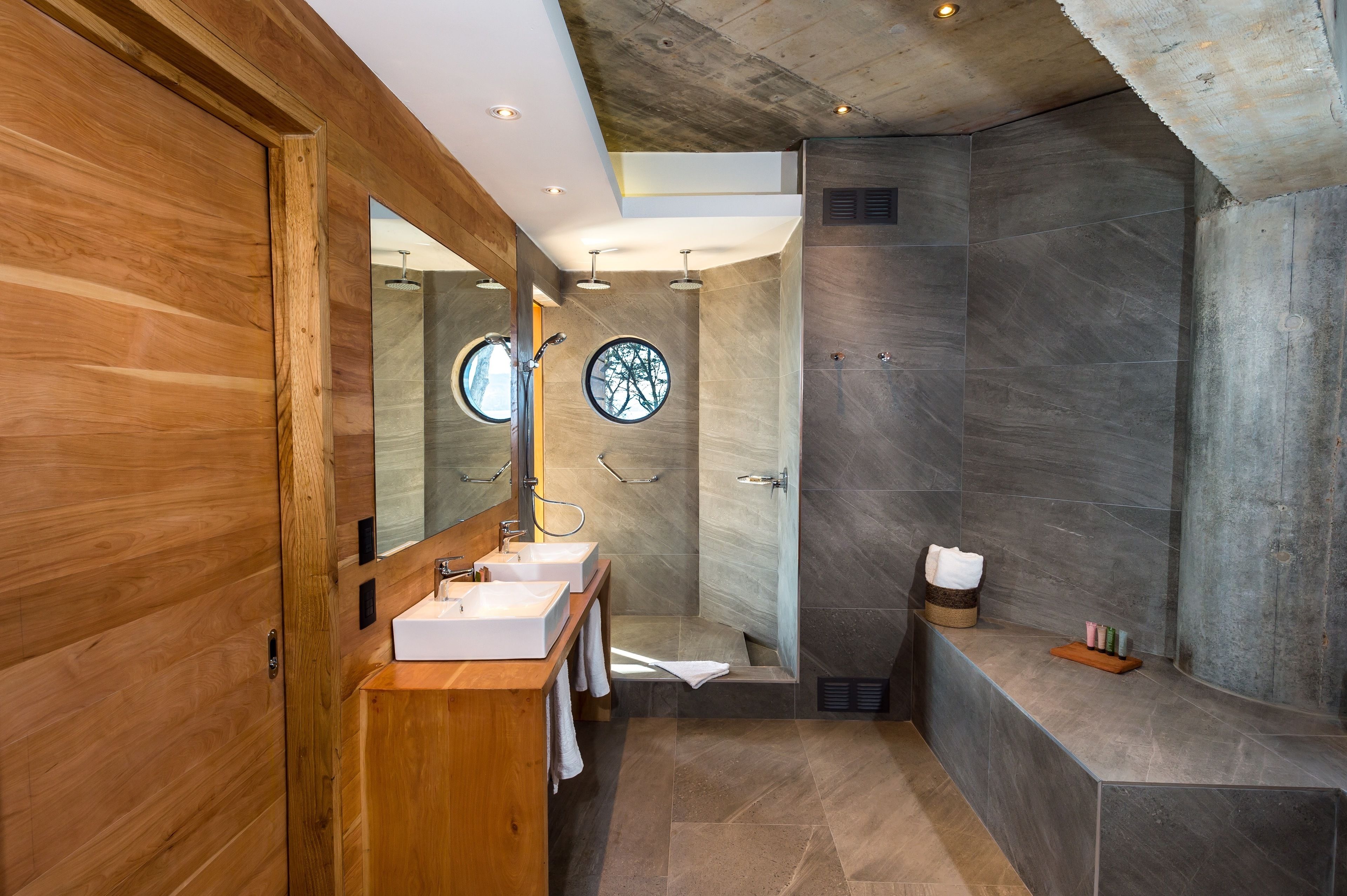 A modern bathroom with a wooden vanity, a round mirror, and a shower with a view of trees through the window.