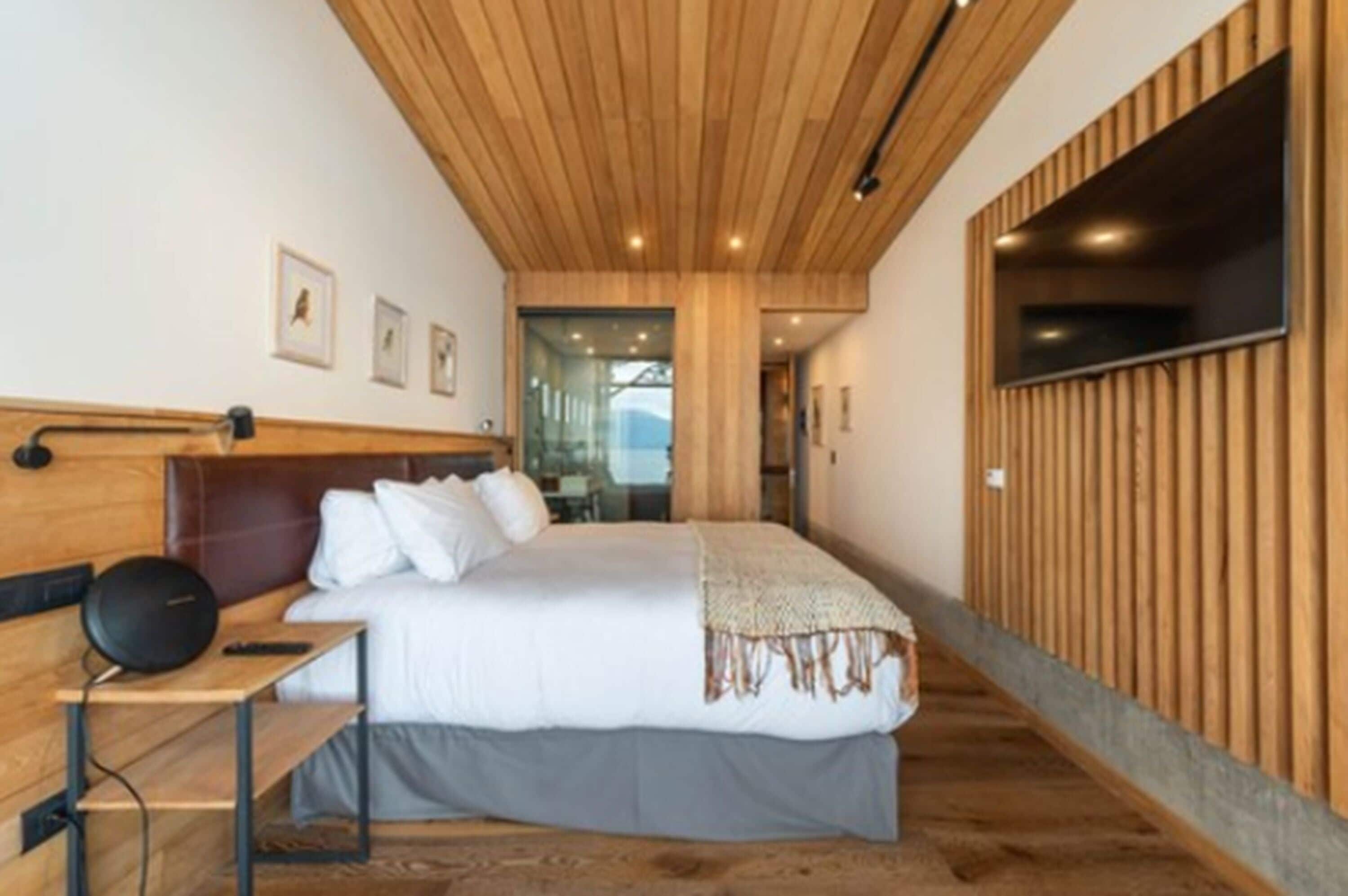 A modern hotel room with a wooden ceiling, a large bed with white bedding, a bedside table with a lamp, and a TV mounted on the wall.