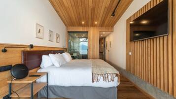 A modern hotel room with a wooden ceiling, a large bed with white bedding, a bedside table with a lamp, and a TV mounted on the wall.