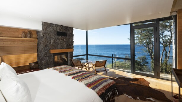 Een slaapkamer met een groot bed, een stenen openhaard, uitzicht op de zee en een balkon met stoelen.