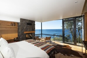Une chambre avec un grand lit, une cheminée en pierre, une vue sur la mer et un balcon avec des chaises.