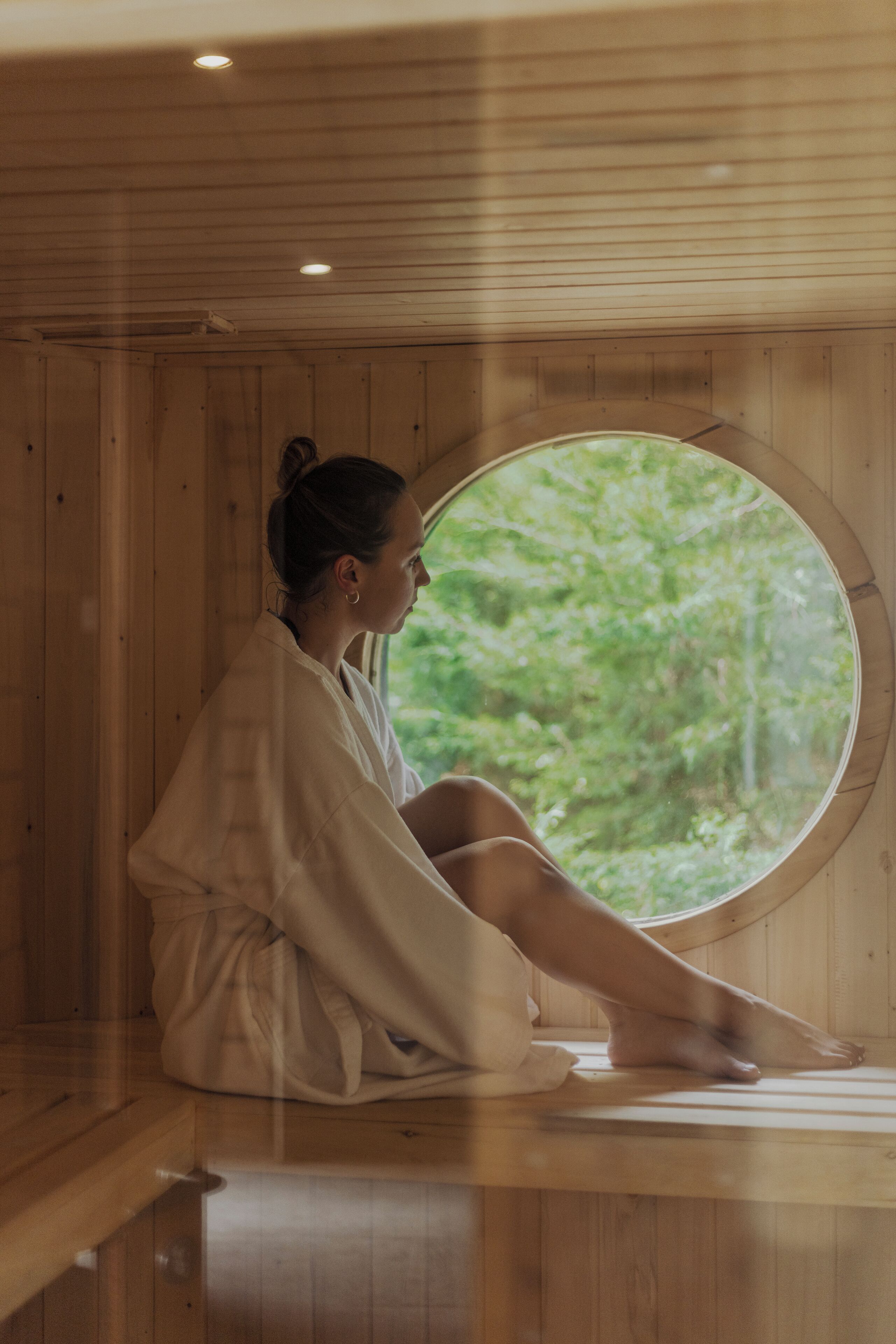 Una persona sentada dentro de un sauna de madera con un amplio ventanal circular.