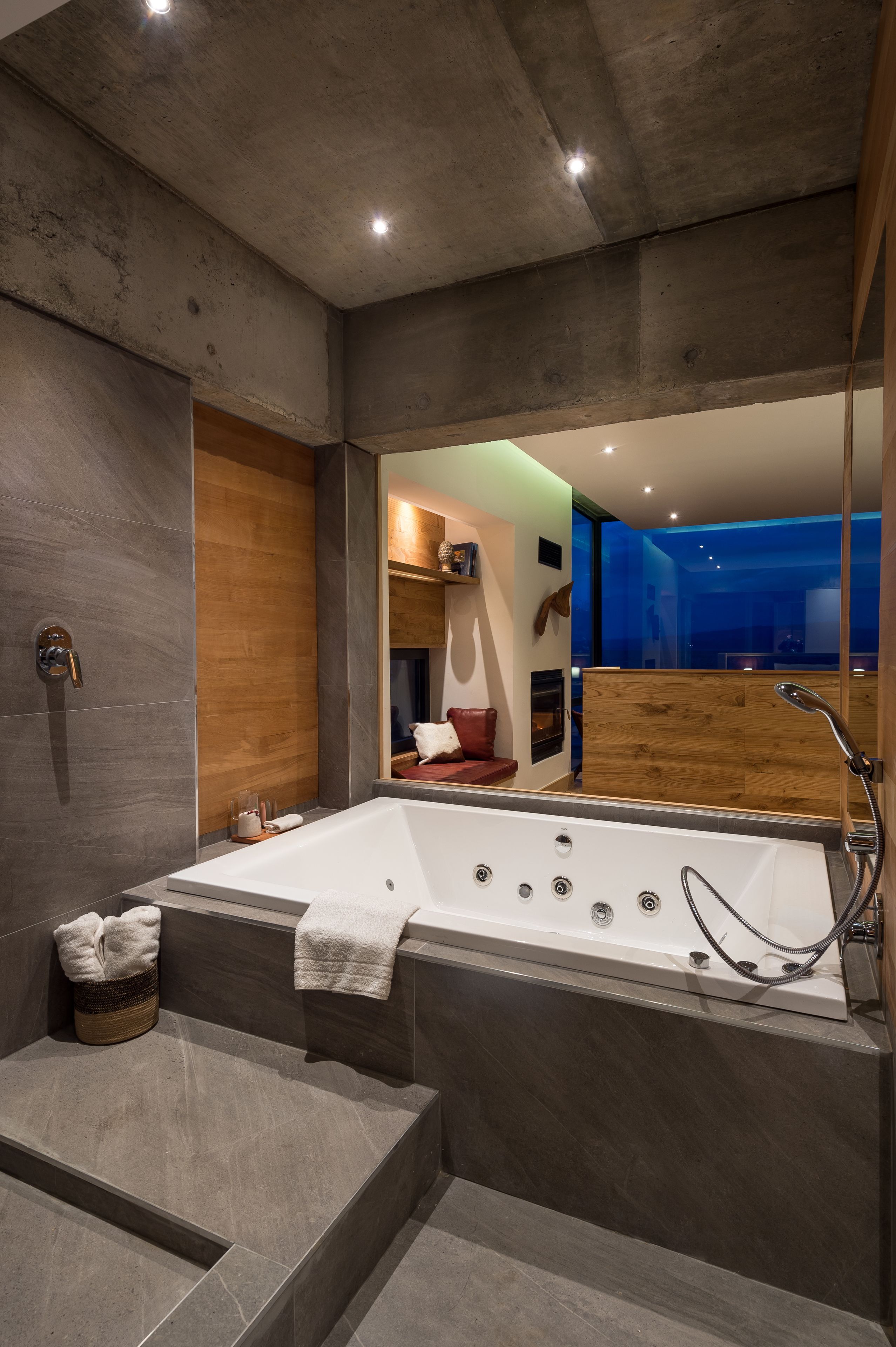 Un baño moderno con una bañera grande, un panel de pared de madera y un espejo que refleja una habitación con un sofá rojo y vistas al exterior.