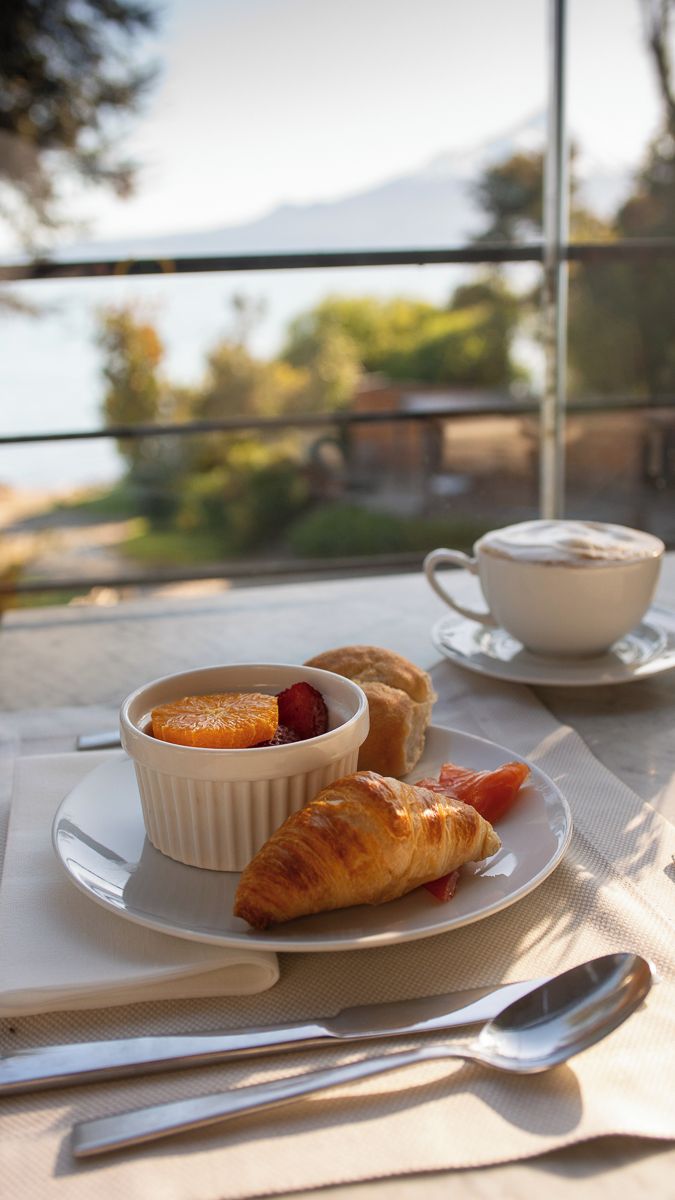A table with a dessert dish containing a fruit, a croissant, and a cup of coffee on a balcony overlooking a scenic view.