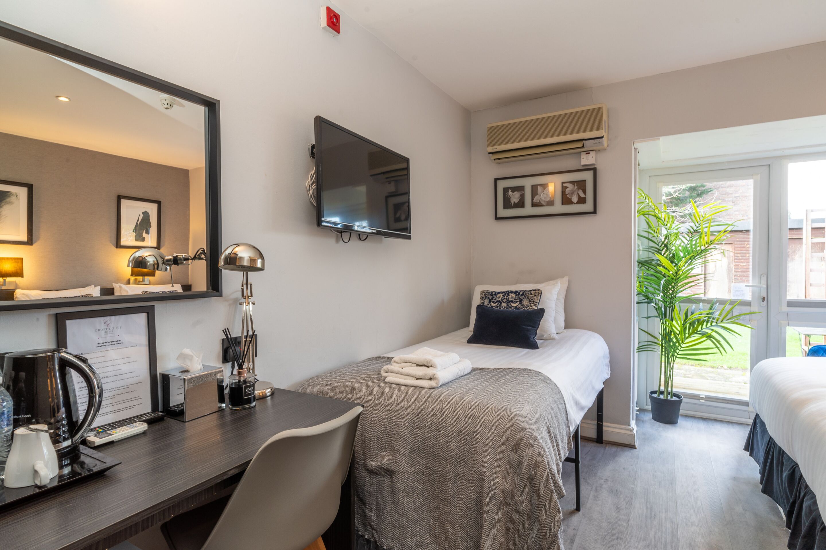 A hotel room with a bed, a desk with a computer, a TV, and a potted plant.