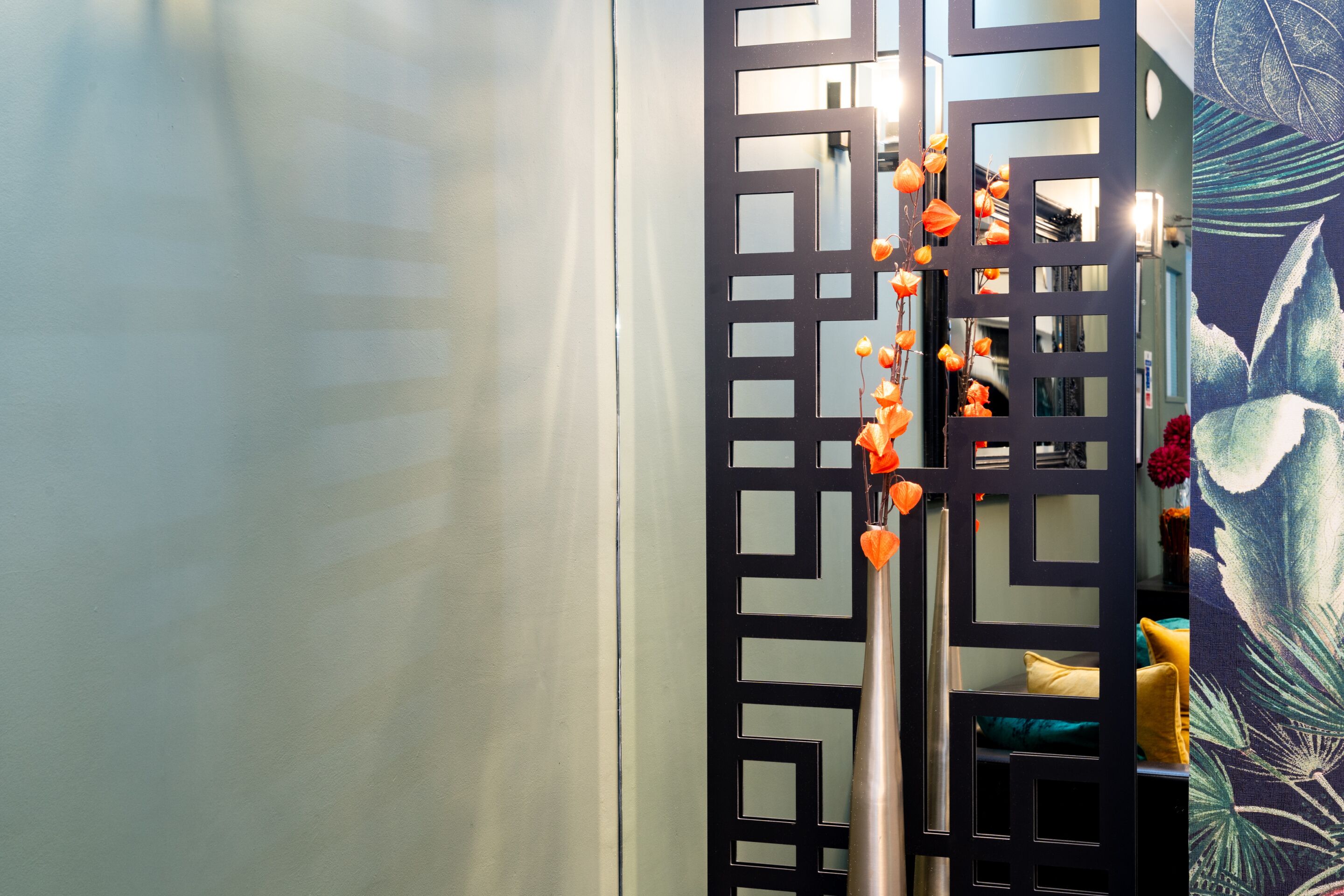 A room with a decorative black partition, a metal stand with orange flowers, and a colorful wall with a leaf pattern.