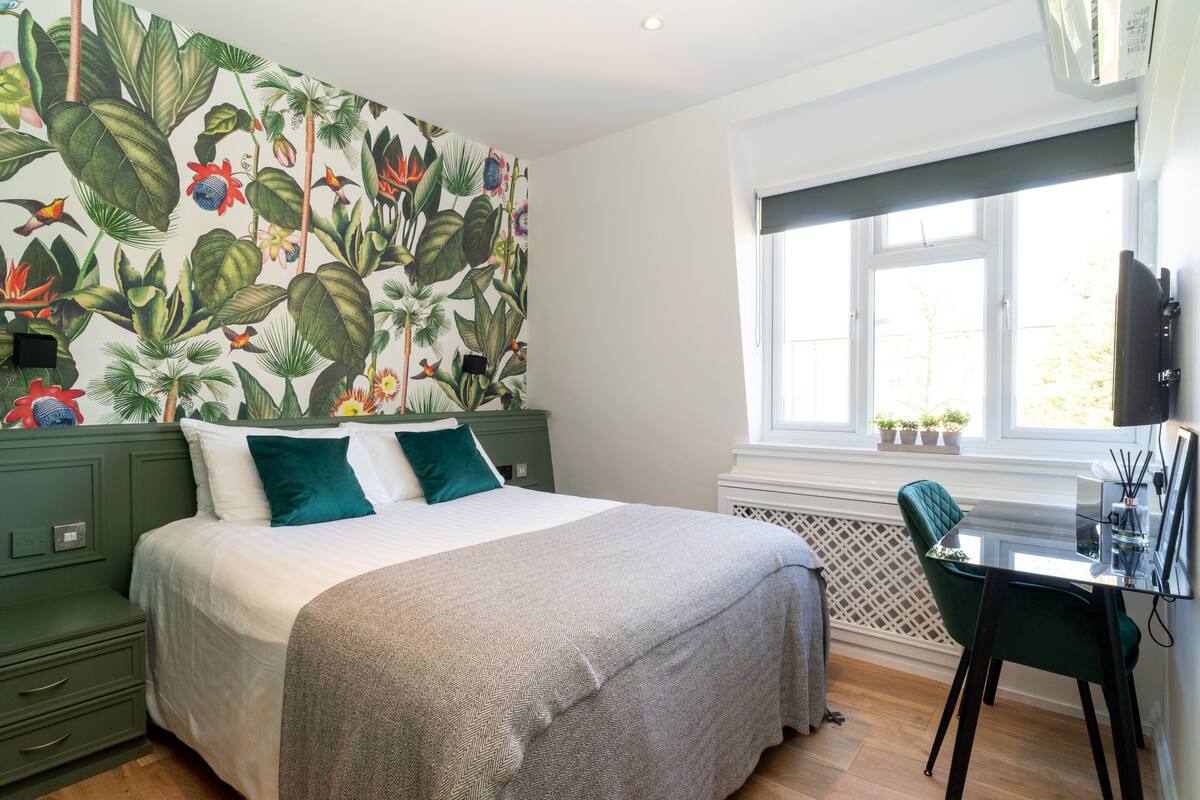 A bedroom with a bed, green nightstands, a desk with a chair, a window, and a wall with a tropical wallpaper.