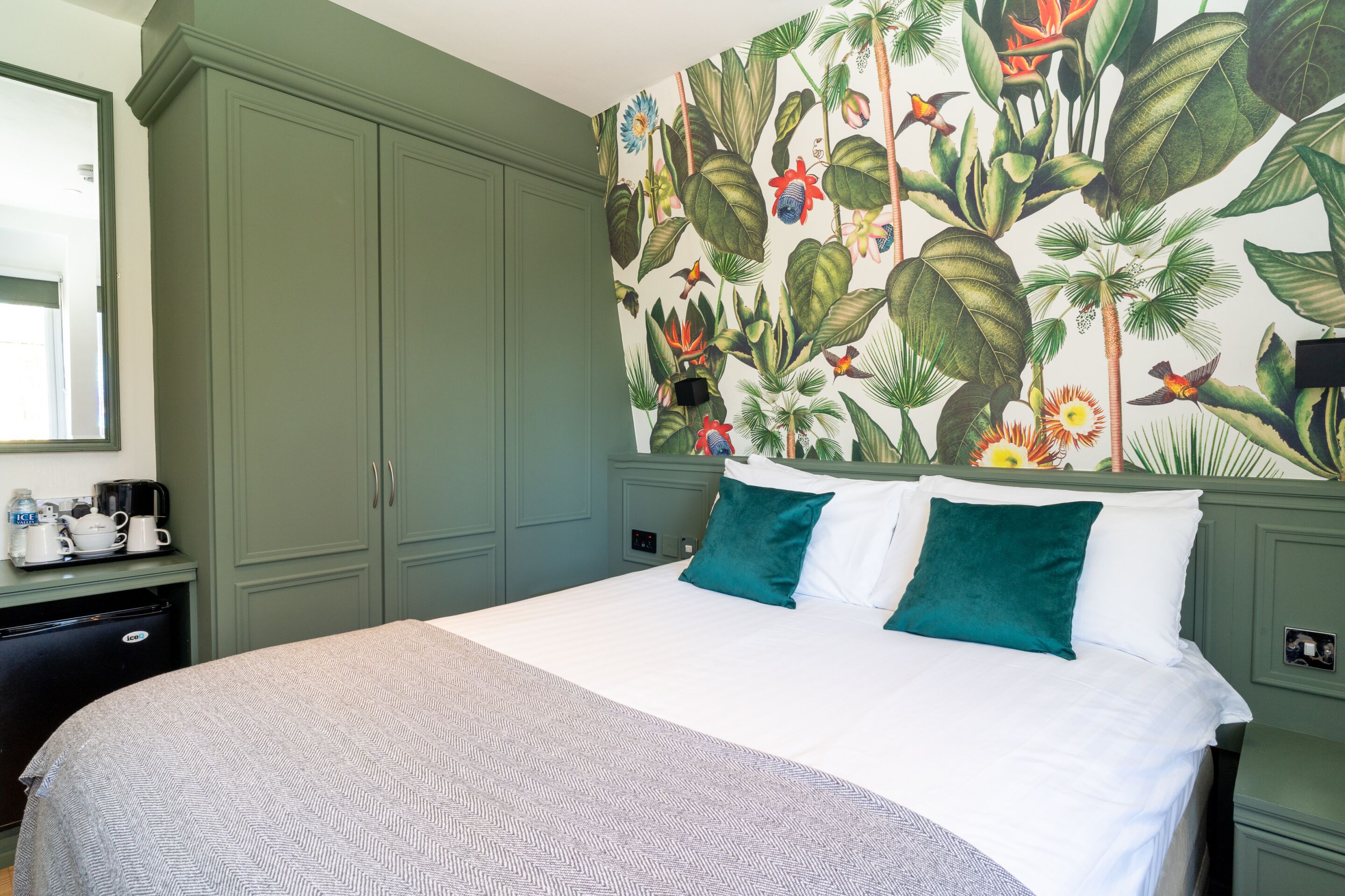 A hotel room with a bed, green cabinets, a desk with a coffee maker, and a tropical-themed wall mural.