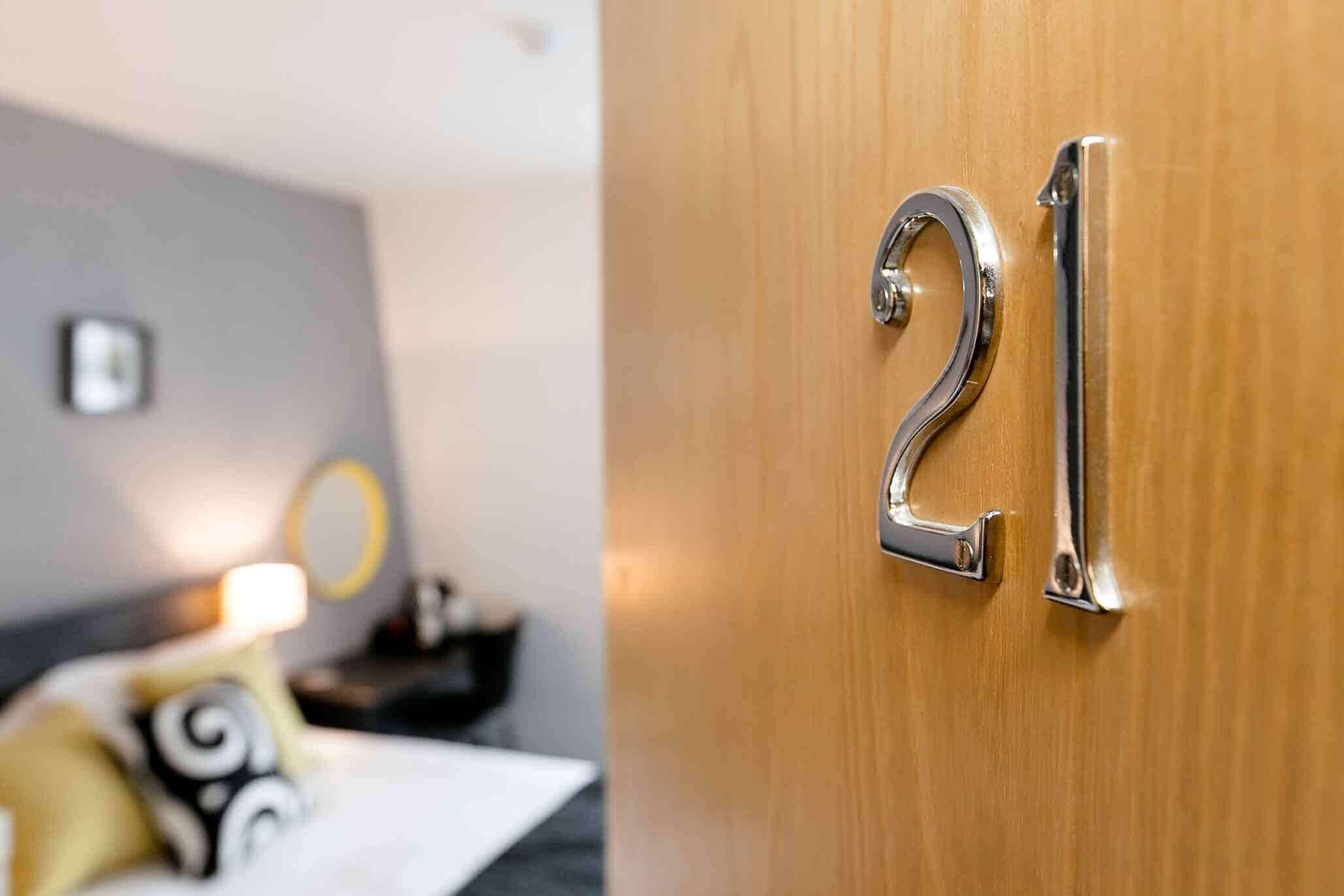 Et hotelværelse med dør nummer 21, en seng med hvid gavl, et natbord og en lampe.