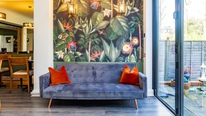 Ein modernes Wohnzimmer mit einem grauen Sofa, zwei orangefarbenen Kissen und einem Wandgemälde mit tropischem Motiv.
