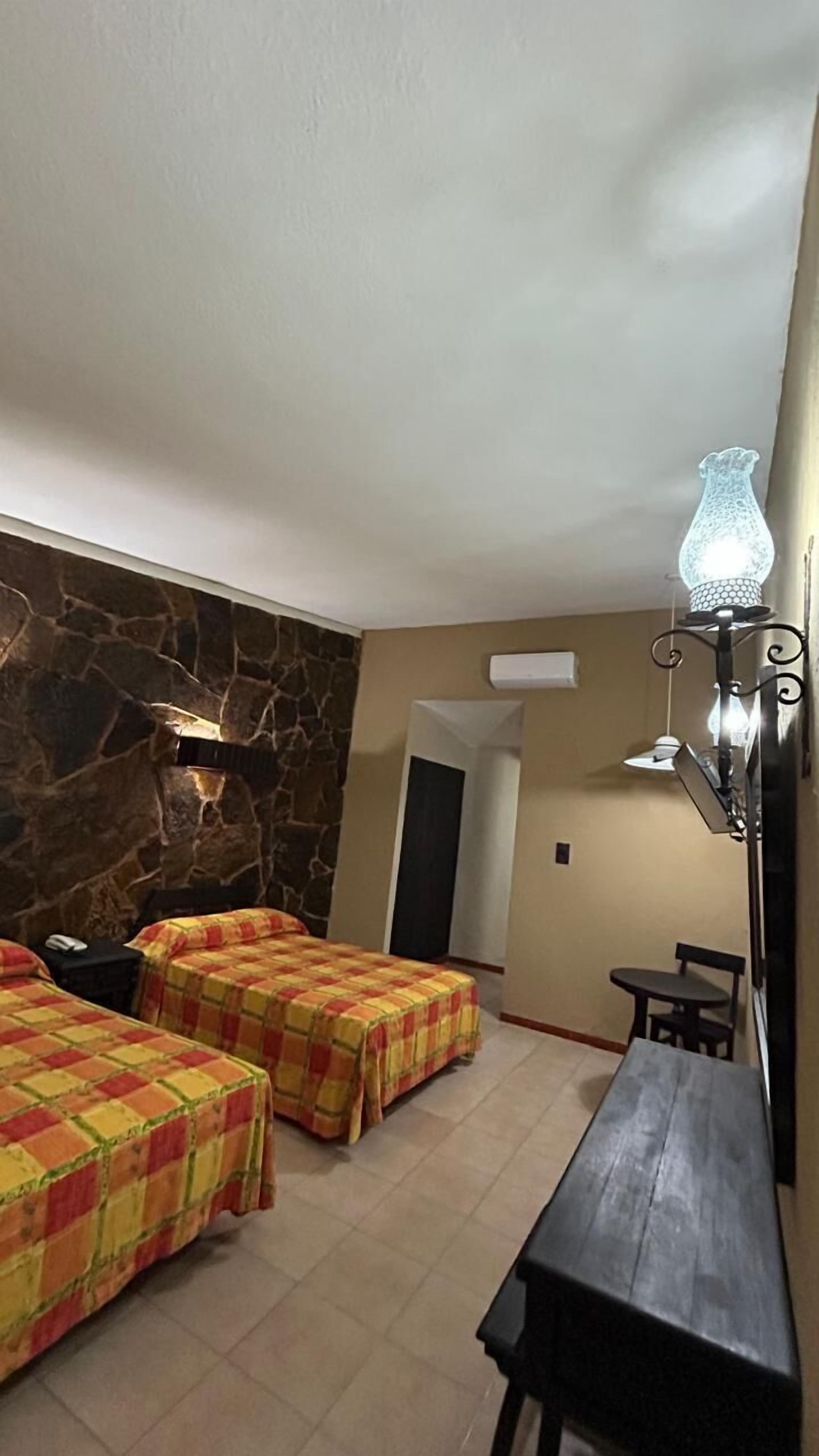 Una habitación con dos camas, una pared de piedra, una puerta, una mesa y una lámpara colgante.