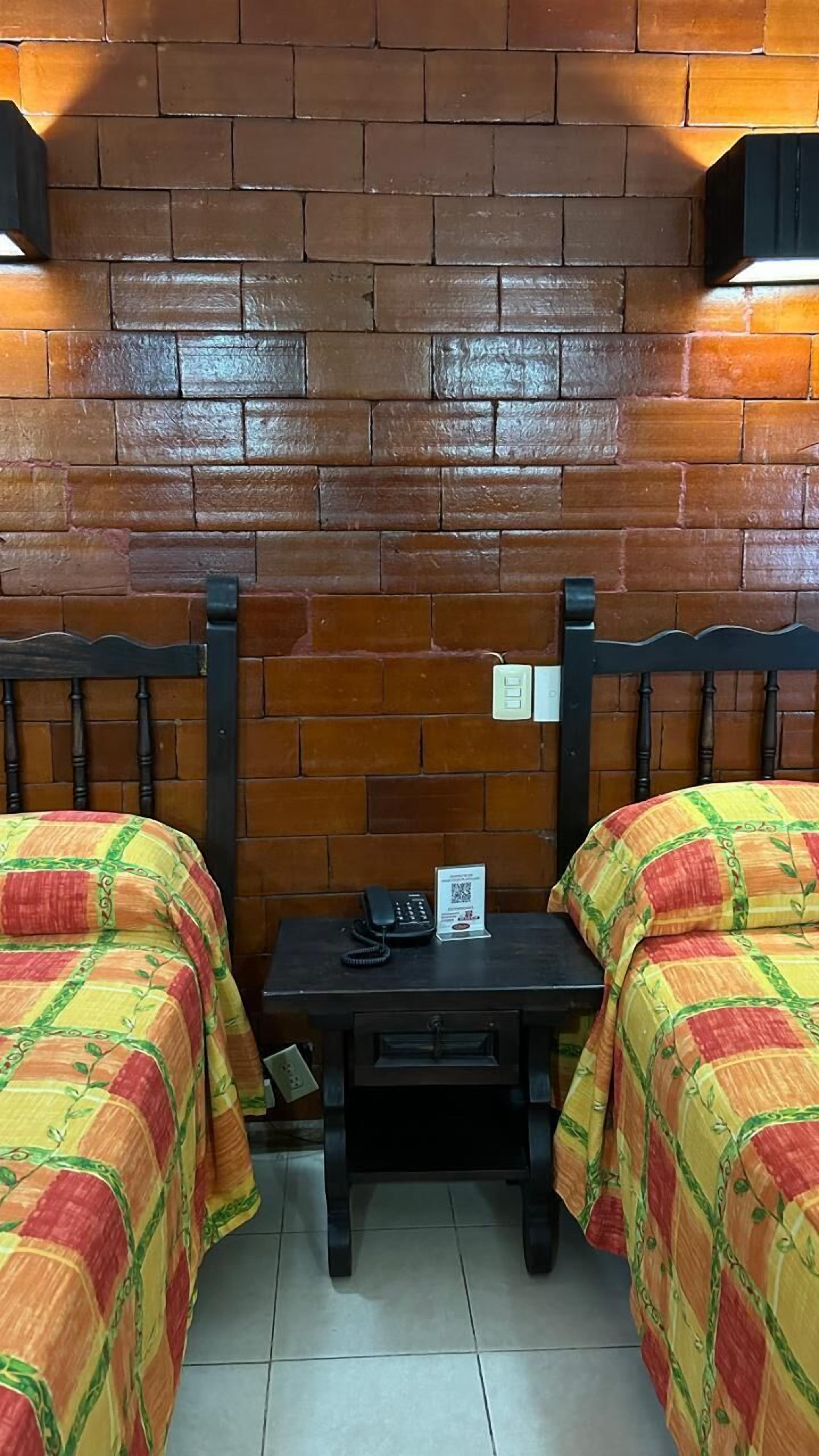 A room with two beds, a small table with a phone, and a brick wall.