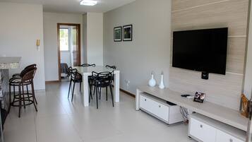 Una sala de estar moderna con un televisor de pantalla plana, una zona de comedor con sillas y una cocina con refrigerador.